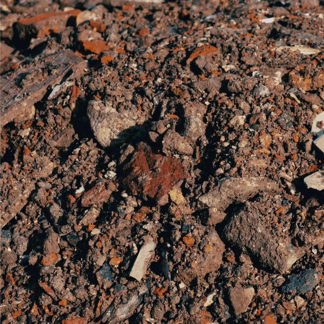 An image of a pile of 6F2 recycled aggregate, typically consisting of crushed concrete, bricks, and asphalt. The aggregate is irregular in shape and size, with pieces varying in size from small pebbles to larger chunks. The colors are a mix of grey, brown, and occasional reddish tones. The pile is unevenly heaped, with some areas higher than others, and it rests on a flat ground, possibly a construction site or recycling facility. The background might be industrial or outdoor, with machinery or open sky visible.